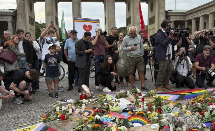 People stand in front of flowers and candles placed at a makeshift memorial in central Berlin on July 26, 2026, a day after a vehicle driven by a man plowed through a crowd of revelers taking part in a nearby parade for sexual minorities, leaving one person dead and about 30 injured. (Kyodo via AP Images)