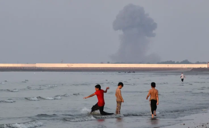 Three boys play in the shallow waters of the Strait of Hormuz, as a plume of smoke rises from an explosion in the background, off Bandar Abbas, Iran, Monday, July 13, 2026. (Razieh Poudat/ISNA via AP)